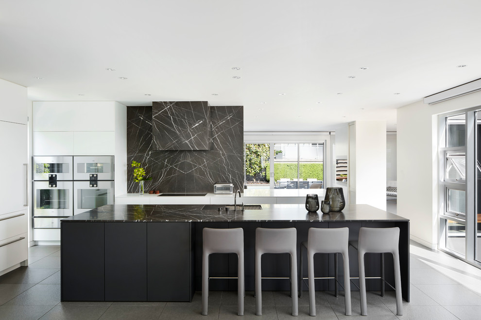 Пример оригинального дизайна: кухня в стиле модернизм с монолитной мойкой, плоскими фасадами, белыми фасадами, черным фартуком, фартуком из каменной плиты, островом, серым полом и черной столешницей