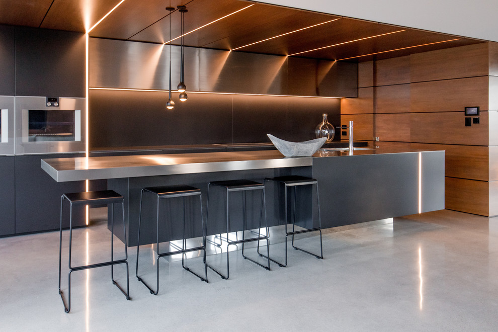 Стильный дизайн: большая параллельная кухня в стиле модернизм с обеденным столом, одинарной мойкой, плоскими фасадами, черными фасадами, столешницей из нержавеющей стали, черным фартуком, бетонным полом, островом и черной столешницей - последний тренд