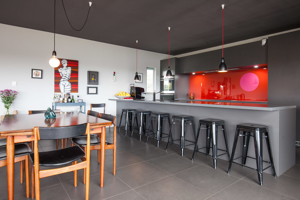 Источник вдохновения для домашнего уюта: параллельная кухня-гостиная в современном стиле с плоскими фасадами, серыми фасадами, красным фартуком, фартуком из стекла, черной техникой и полуостровом