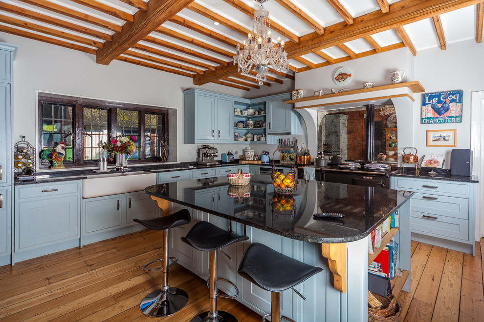 This is an example of a farmhouse l-shaped kitchen in Kent with a belfast sink, shaker cabinets, blue cabinets, black appliances, medium hardwood flooring, an island, brown floors and black worktops.
