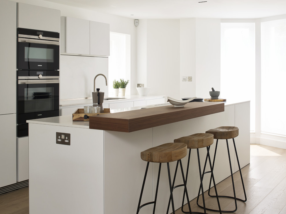 Photo of a contemporary galley kitchen in London with flat-panel cabinets, white cabinets, black appliances, light hardwood flooring and an island.