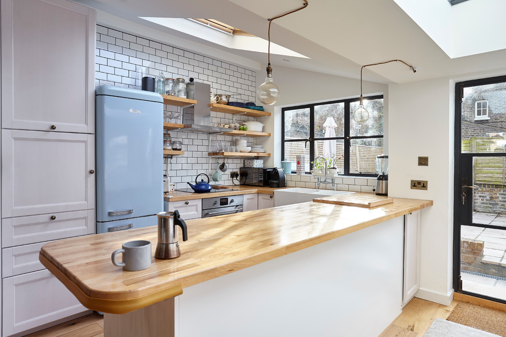 Example of a small country u-shaped light wood floor kitchen design in London with a farmhouse sink, recessed-panel cabinets, white cabinets, wood countertops, white backsplash, subway tile backsplash, colored appliances and a peninsula
