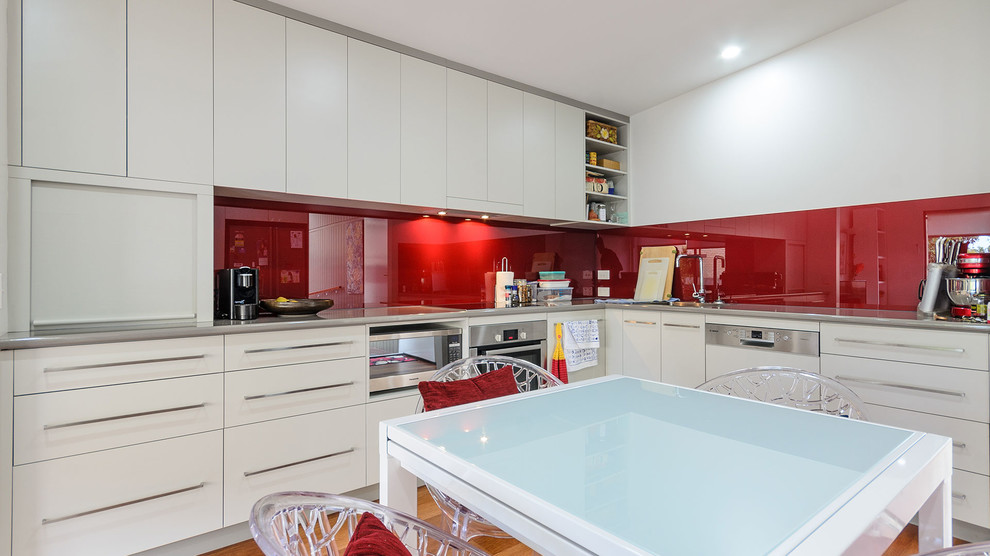 Kleine Moderne Wohnküche ohne Insel in L-Form mit Waschbecken, weißen Schränken, Laminat-Arbeitsplatte, Küchenrückwand in Rot, Glasrückwand, Küchengeräten aus Edelstahl und hellem Holzboden in Brisbane