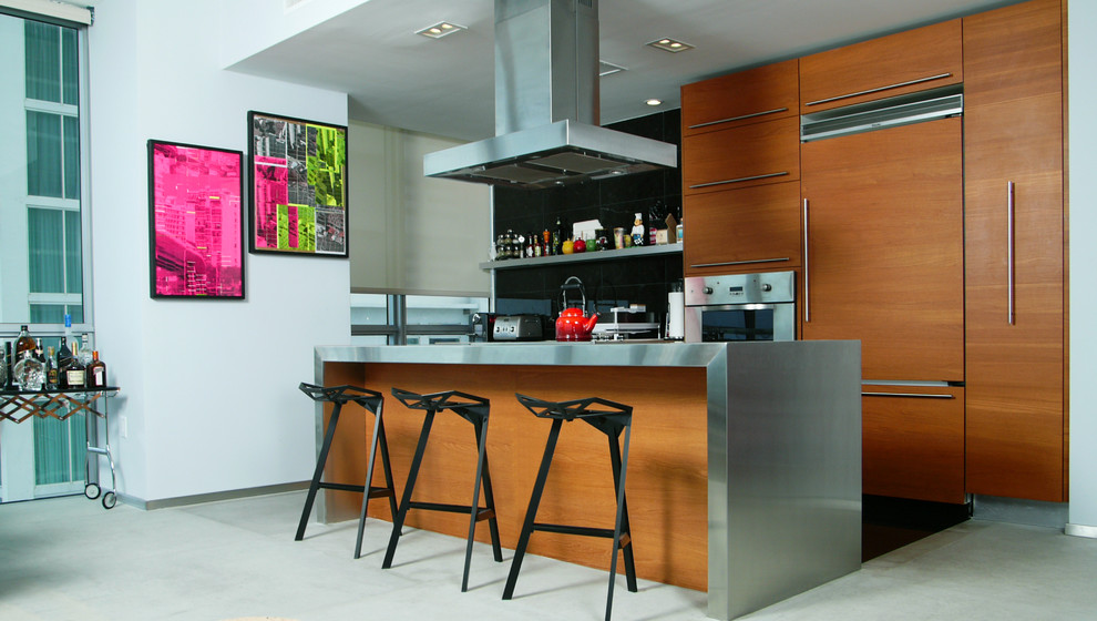 Zweizeilige Moderne Küche mit flächenbündigen Schrankfronten, hellbraunen Holzschränken, Edelstahl-Arbeitsplatte, Küchenrückwand in Schwarz, Elektrogeräten mit Frontblende, Betonboden und Kücheninsel in Miami