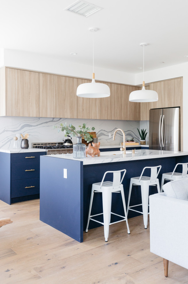 Design ideas for a large country l-shaped open plan kitchen in Los Angeles with a belfast sink, flat-panel cabinets, blue cabinets, white splashback, porcelain splashback, stainless steel appliances, light hardwood flooring, an island, beige floors and white worktops.