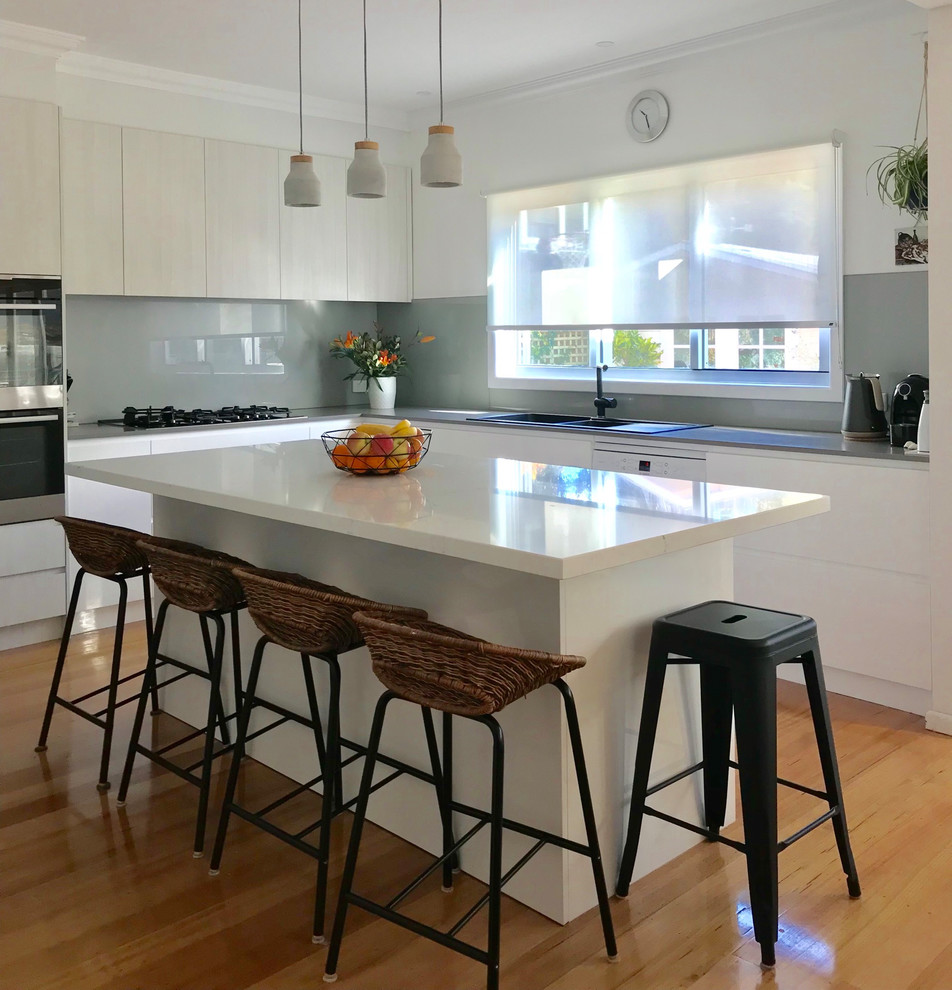 This is an example of a medium sized contemporary l-shaped kitchen/diner in Melbourne with a double-bowl sink, white cabinets, marble worktops, metallic splashback, glass sheet splashback, black appliances, light hardwood flooring, an island, brown floors and grey worktops.
