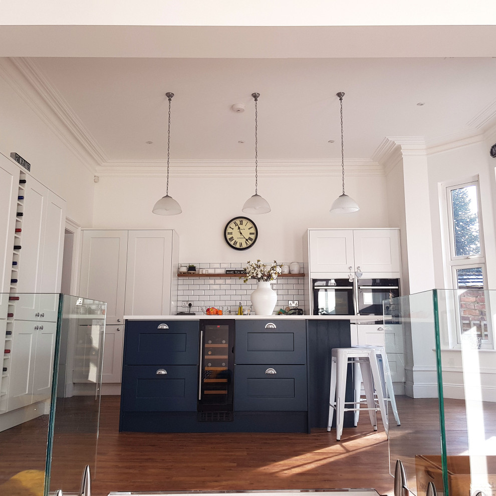 This is an example of a medium sized traditional l-shaped open plan kitchen in Manchester with a belfast sink, recessed-panel cabinets, blue cabinets, quartz worktops, white splashback, metro tiled splashback, integrated appliances, medium hardwood flooring, an island, brown floors and white worktops.