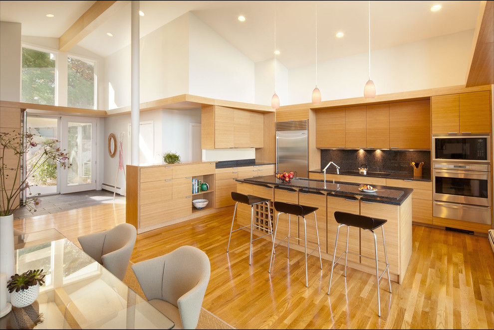 Modelo de cocina comedor actual con armarios con paneles lisos, puertas de armario de madera clara, salpicadero negro y electrodomésticos de acero inoxidable