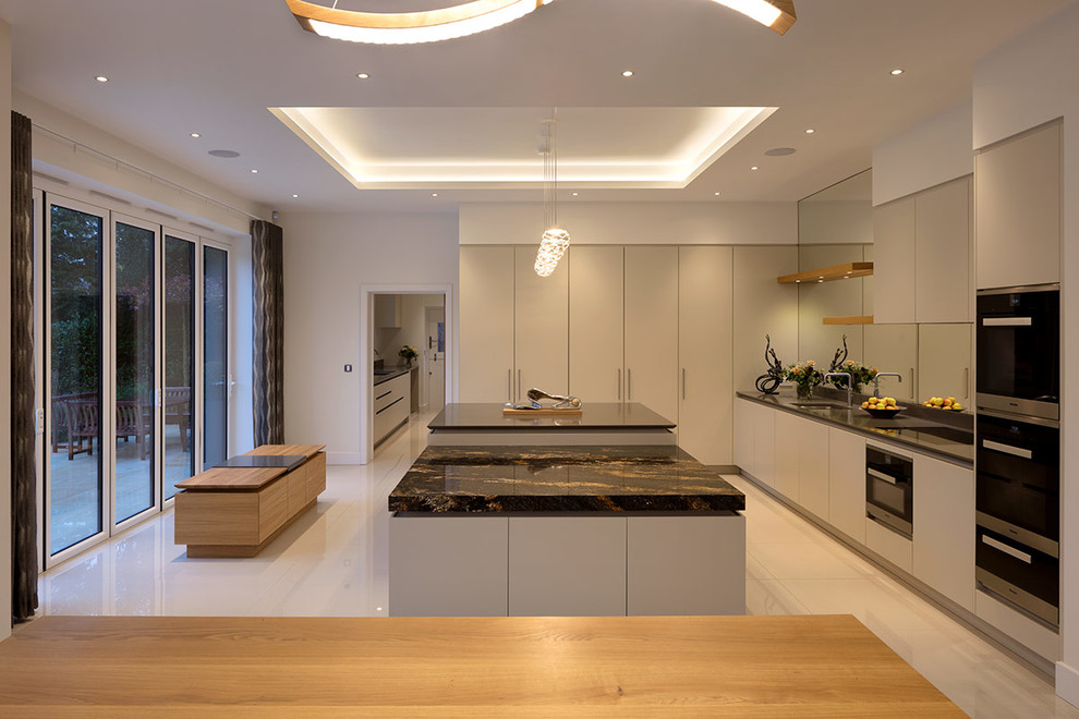 Design ideas for an expansive contemporary l-shaped open plan kitchen in London with flat-panel cabinets, beige cabinets, granite worktops, metallic splashback, mirror splashback, stainless steel appliances, an island, white floors and black worktops.