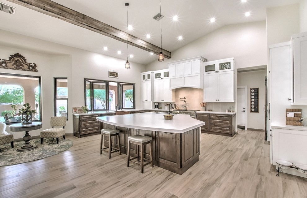 Photo of a large country l-shaped kitchen/diner in Phoenix with a belfast sink, raised-panel cabinets, brown cabinets, engineered stone countertops, white splashback, porcelain splashback, stainless steel appliances, porcelain flooring, an island, brown floors and white worktops.