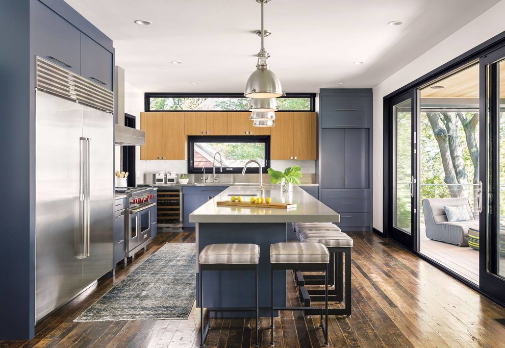 Modern inredning av ett kök, med en enkel diskho, släta luckor, blå skåp, bänkskiva i rostfritt stål, stänkskydd med metallisk yta, rostfria vitvaror, mörkt trägolv, en köksö och brunt golv