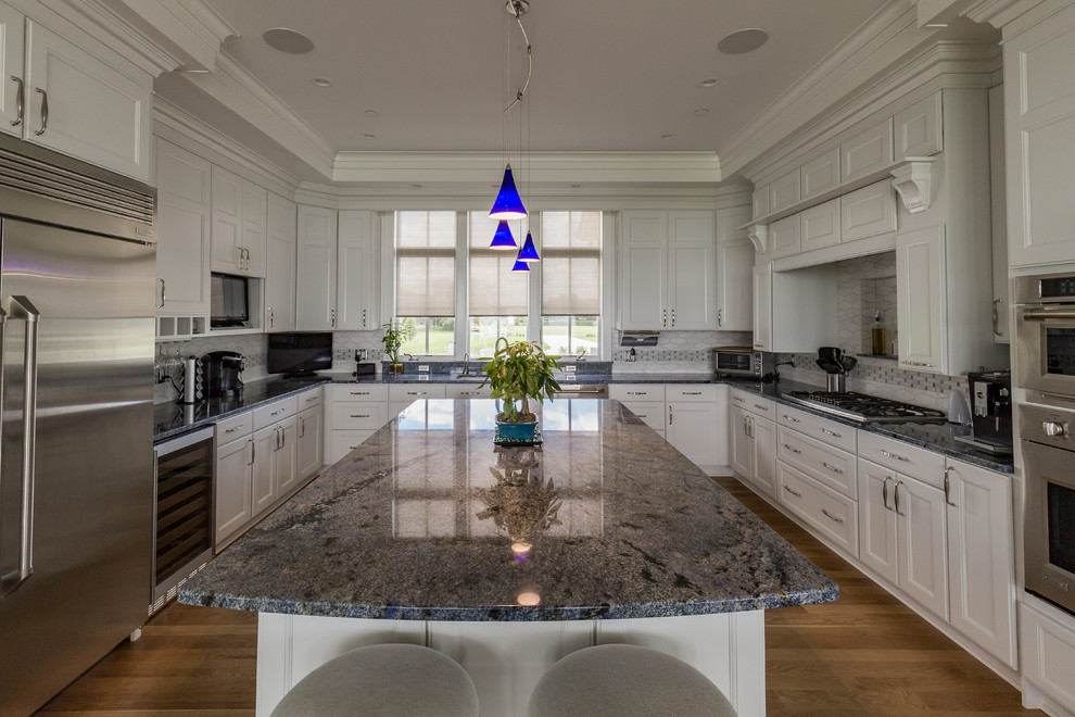 Idéer för att renovera ett mycket stort amerikanskt blå blått kök, med en dubbel diskho, luckor med infälld panel, vita skåp, granitbänkskiva, grått stänkskydd, stänkskydd i mosaik, rostfria vitvaror, mellanmörkt trägolv, en köksö och brunt golv