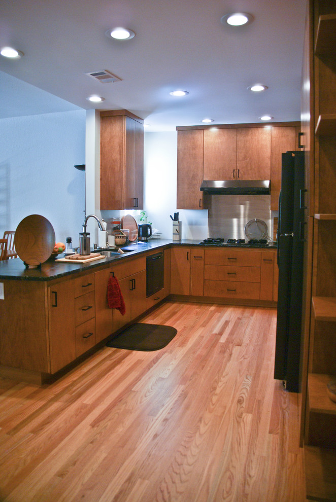 This is an example of a small contemporary u-shaped kitchen/diner in Sacramento with a built-in sink, flat-panel cabinets, medium wood cabinets, soapstone worktops, black appliances, medium hardwood flooring and a breakfast bar.