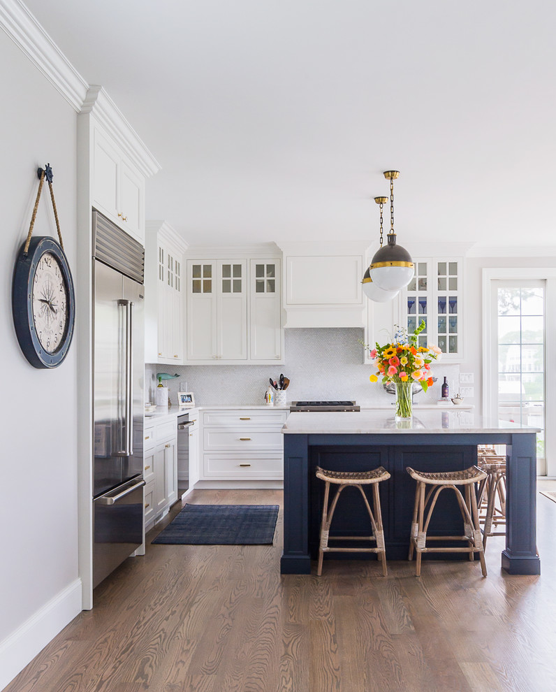 Modelo de cocina clásica renovada, grande, con armarios estilo shaker, puertas de armario blancas, salpicadero blanco, electrodomésticos de acero inoxidable, suelo de madera en tonos medios, una isla, suelo marrón y encimeras blancas