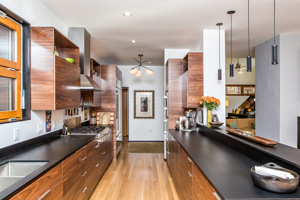 Large midcentury galley kitchen/diner in Raleigh with a submerged sink, flat-panel cabinets, dark wood cabinets, laminate countertops, black splashback, stainless steel appliances, medium hardwood flooring, an island, brown floors and black worktops.