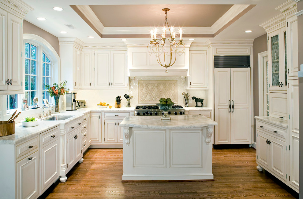 Klassisk inredning av ett l-kök, med en undermonterad diskho, luckor med upphöjd panel, vita skåp, beige stänkskydd, integrerade vitvaror, mellanmörkt trägolv och en köksö