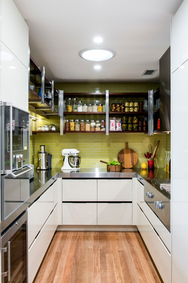 Foto på ett avskilt, litet funkis u-kök, med släta luckor, vita skåp, bänkskiva i rostfritt stål, grönt stänkskydd och mellanmörkt trägolv