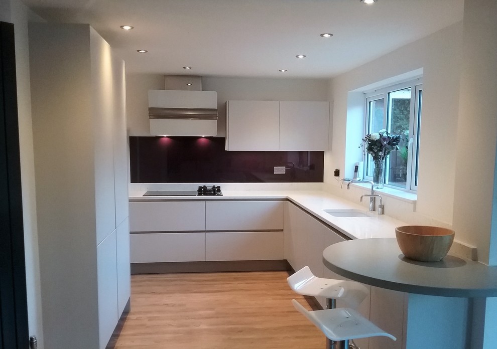 Photo of a contemporary l-shaped kitchen/diner in West Midlands with a submerged sink, flat-panel cabinets, beige cabinets, quartz worktops, red splashback, glass sheet splashback and black appliances.