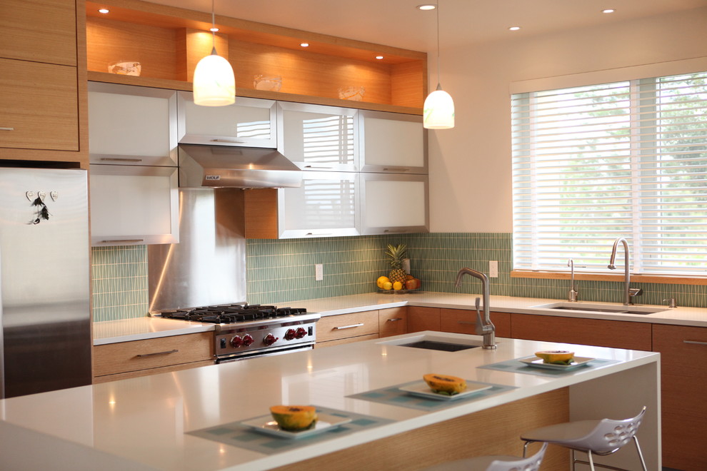 Inredning av ett modernt l-kök, med en undermonterad diskho, luckor med glaspanel, skåp i rostfritt stål, blått stänkskydd, stänkskydd i glaskakel och rostfria vitvaror