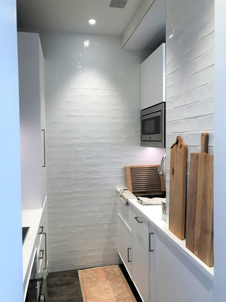 Photo of a small contemporary galley enclosed kitchen in New York with a submerged sink, recessed-panel cabinets, white cabinets, engineered stone countertops, white splashback, cement tile splashback, white appliances, dark hardwood flooring, an island, brown floors and white worktops.