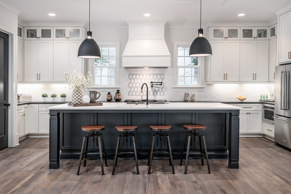 This is an example of a traditional u-shaped kitchen in Charlotte with shaker cabinets, white cabinets, white splashback, stainless steel appliances, medium hardwood flooring, an island, brown floors and white worktops.