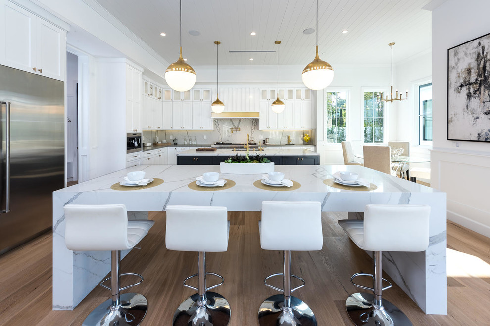 This is an example of a classic kitchen/diner in Los Angeles with a submerged sink, shaker cabinets, white cabinets, white splashback, stainless steel appliances, light hardwood flooring, multiple islands and beige floors.
