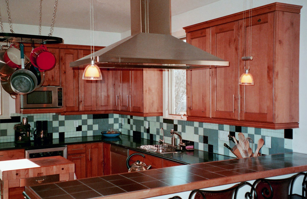 Bild på ett avskilt, mellanstort u-kök, med en nedsänkt diskho, luckor med infälld panel, skåp i mellenmörkt trä, kaklad bänkskiva, grönt stänkskydd, stänkskydd i glaskakel, rostfria vitvaror och en köksö