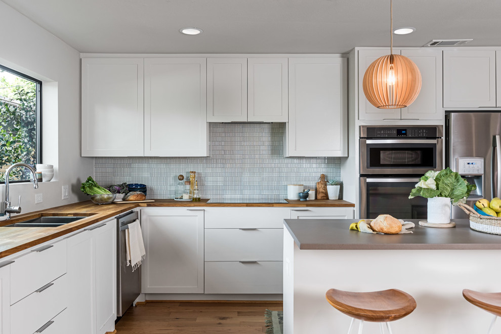 This is an example of a traditional l-shaped kitchen in Houston with a double-bowl sink, shaker cabinets, white cabinets, grey splashback, stainless steel appliances, light hardwood flooring, an island, beige floors and wood worktops.