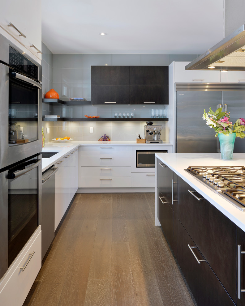 Bild på ett funkis l-kök, med en undermonterad diskho, släta luckor, vita skåp, grått stänkskydd, rostfria vitvaror, mellanmörkt trägolv, en köksö och glaspanel som stänkskydd