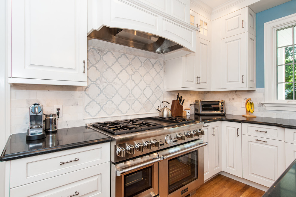 Example of a mid-sized transitional l-shaped medium tone wood floor enclosed kitchen design in DC Metro with an undermount sink, raised-panel cabinets, white cabinets, granite countertops, white backsplash, stone tile backsplash, stainless steel appliances and an island