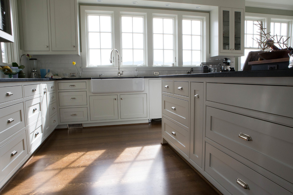 Inredning av ett lantligt stort grå grått kök, med en rustik diskho, luckor med profilerade fronter, vita skåp, stänkskydd i tunnelbanekakel, rostfria vitvaror, mellanmörkt trägolv, en köksö och brunt golv
