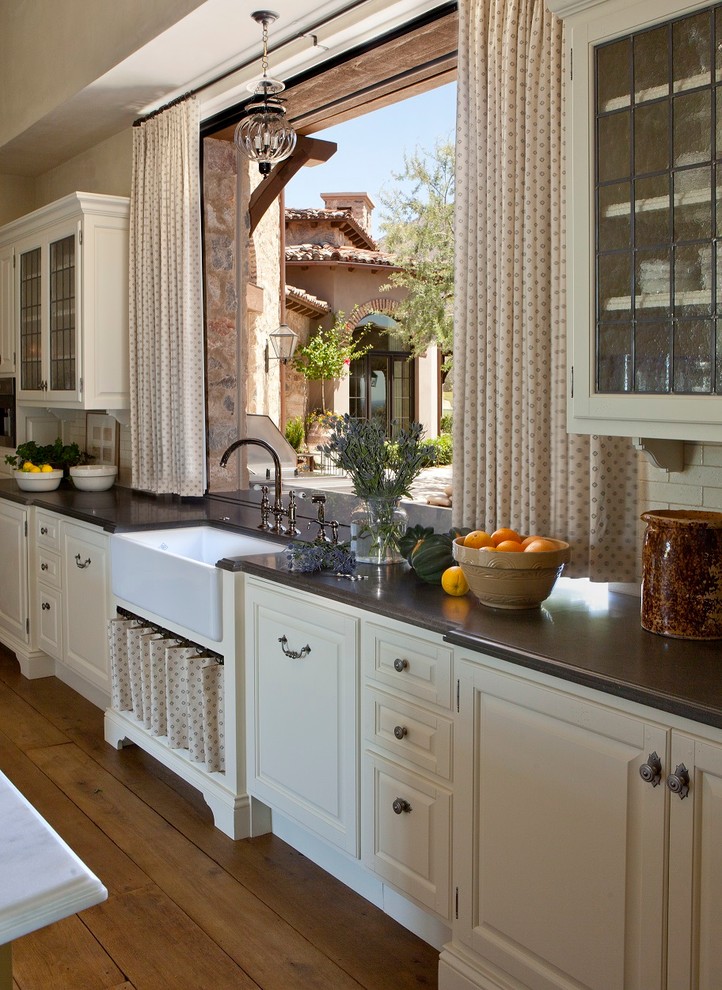 Eat-in kitchen - huge mediterranean l-shaped medium tone wood floor and brown floor eat-in kitchen idea in Phoenix with glass-front cabinets, a farmhouse sink, white cabinets, solid surface countertops, white backsplash, subway tile backsplash, stainless steel appliances and an island