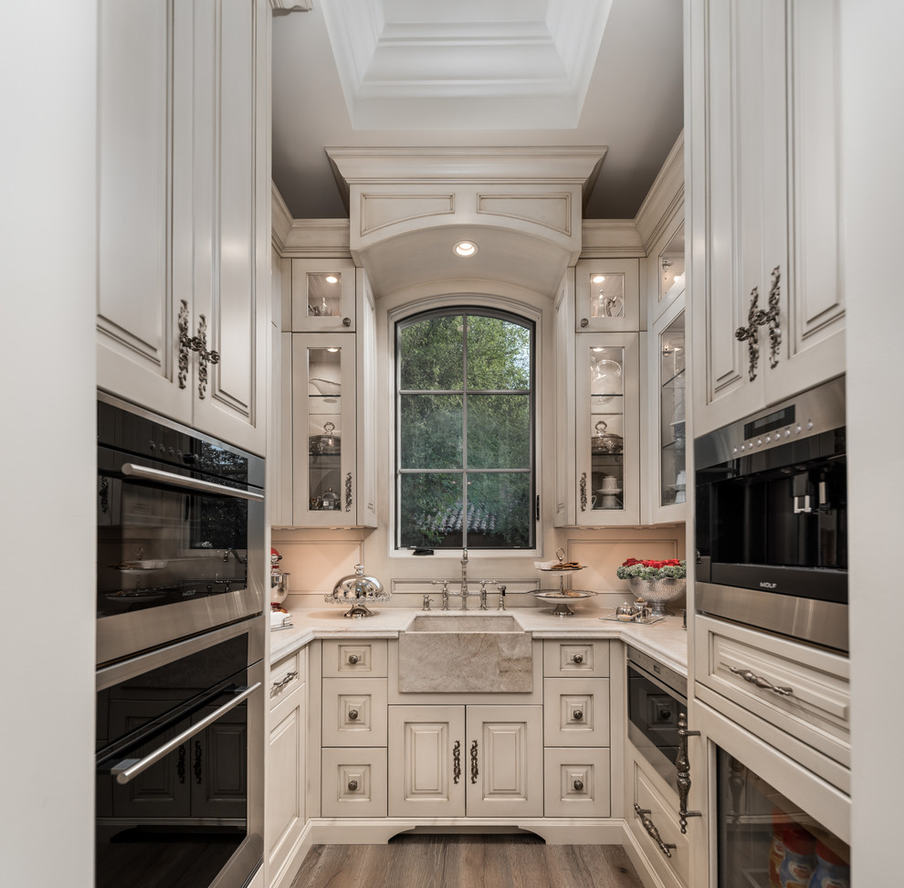 Photo of an expansive mediterranean u-shaped enclosed kitchen in Phoenix with a belfast sink, white splashback, stainless steel appliances, medium hardwood flooring, brown floors, raised-panel cabinets, beige cabinets and white worktops.