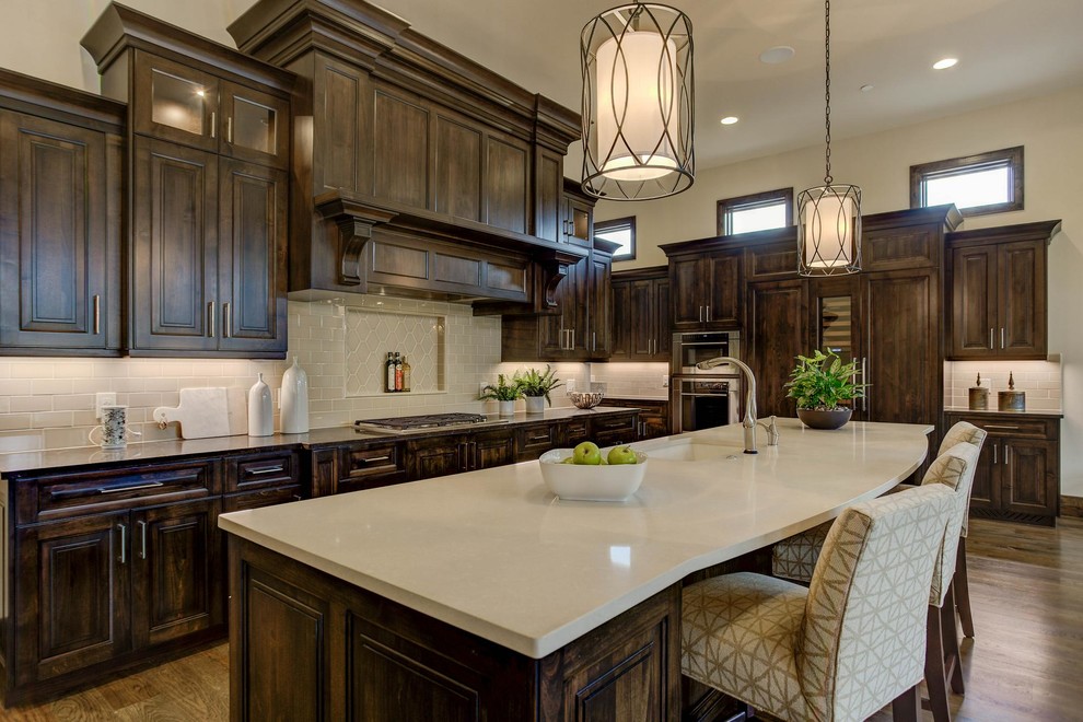 This is an example of a large mediterranean l-shaped open plan kitchen in Denver with a submerged sink, beaded cabinets, dark wood cabinets, engineered stone countertops, beige splashback, metro tiled splashback, integrated appliances, medium hardwood flooring, an island, brown floors and beige worktops.
