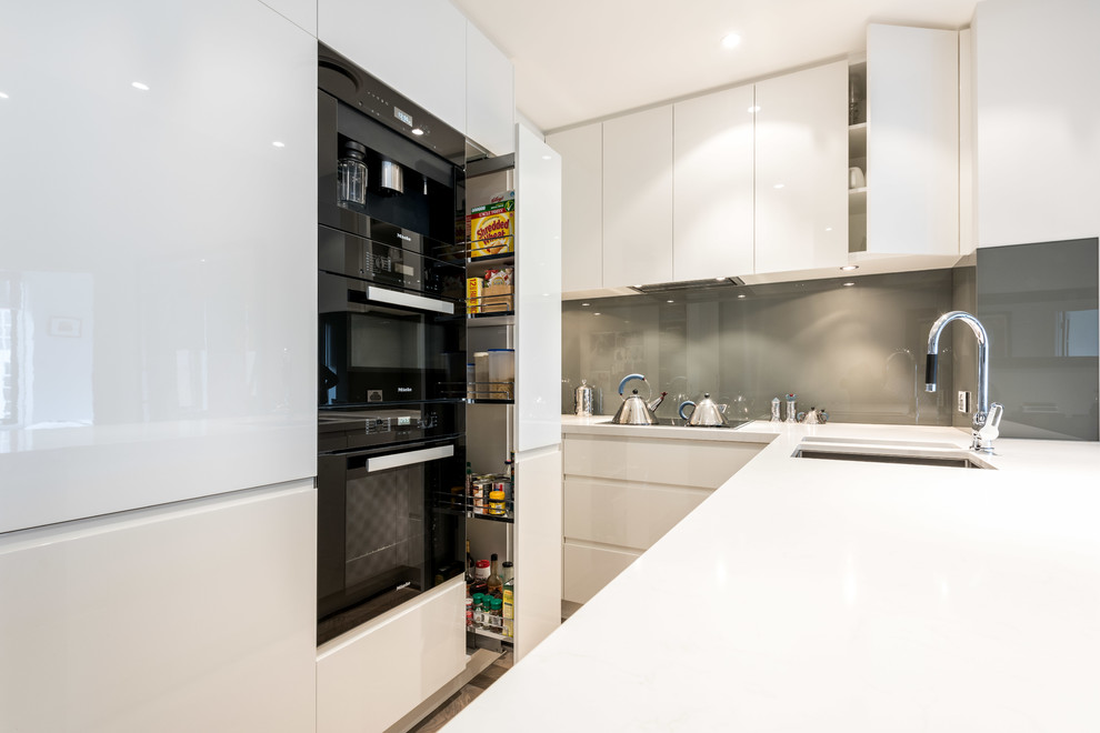 Kleine Moderne Wohnküche ohne Insel in U-Form mit Unterbauwaschbecken, flächenbündigen Schrankfronten, weißen Schränken, Quarzwerkstein-Arbeitsplatte, Küchenrückwand in Grün, Glasrückwand und schwarzen Elektrogeräten in Melbourne