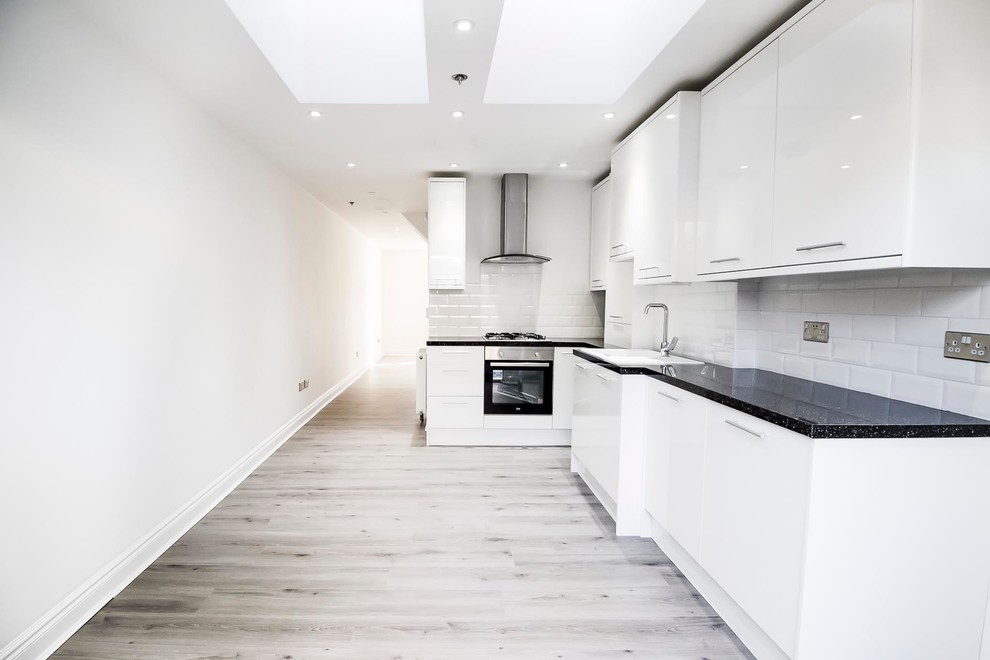 This is an example of a medium sized bohemian l-shaped kitchen/diner in London with flat-panel cabinets, white cabinets, granite worktops, white splashback, ceramic splashback, laminate floors, black appliances and grey floors.
