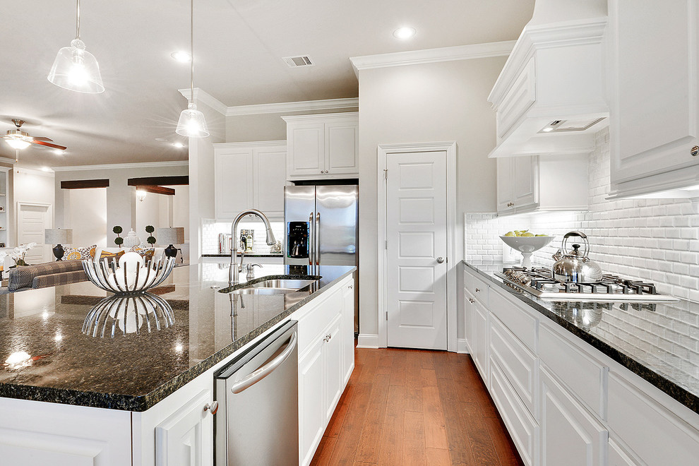 Inspiration for a l-shaped light wood floor and brown floor open concept kitchen remodel in New Orleans with a drop-in sink, raised-panel cabinets, white cabinets, granite countertops, white backsplash, subway tile backsplash, stainless steel appliances, an island and black countertops