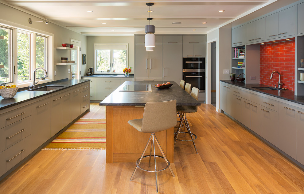 This is an example of a contemporary kitchen in Minneapolis with a submerged sink, flat-panel cabinets, grey cabinets, red splashback, black appliances, light hardwood flooring, an island and metro tiled splashback.