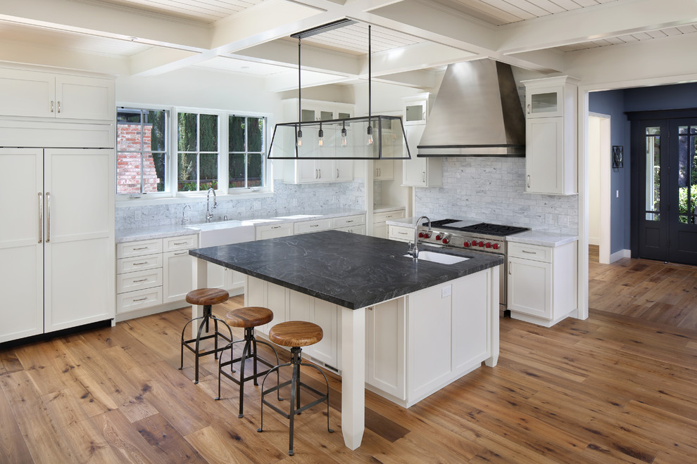 Design ideas for a large farmhouse l-shaped kitchen in San Francisco with a belfast sink, shaker cabinets, white cabinets, soapstone worktops, grey splashback, stainless steel appliances, medium hardwood flooring, an island, stone tiled splashback and brown floors.