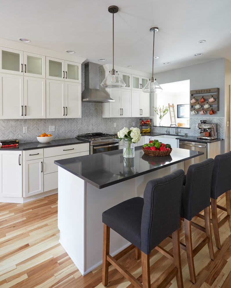 This is an example of a medium sized country l-shaped open plan kitchen in Milwaukee with a belfast sink, recessed-panel cabinets, white cabinets, engineered stone countertops, grey splashback, marble splashback, stainless steel appliances, medium hardwood flooring, an island and brown floors.