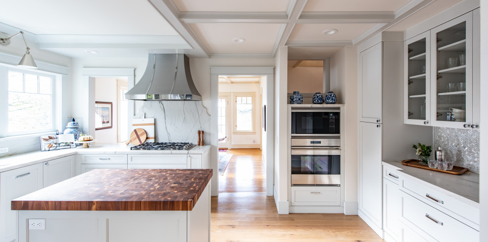 Bild på ett stort lantligt grå grått kök, med vita skåp, grått stänkskydd, stänkskydd i marmor, rostfria vitvaror, ljust trägolv, en köksö och brunt golv