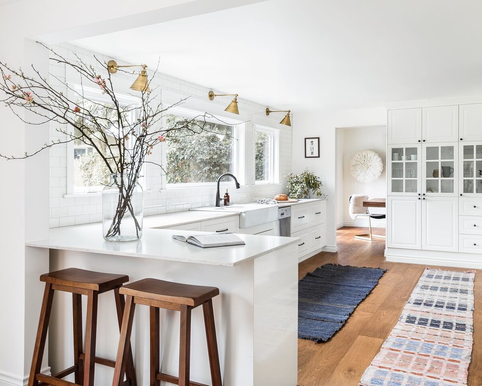 Example of a large country l-shaped medium tone wood floor and brown floor enclosed kitchen design in Seattle with a farmhouse sink, raised-panel cabinets, white cabinets, white backsplash, subway tile backsplash, stainless steel appliances, a peninsula and quartzite countertops