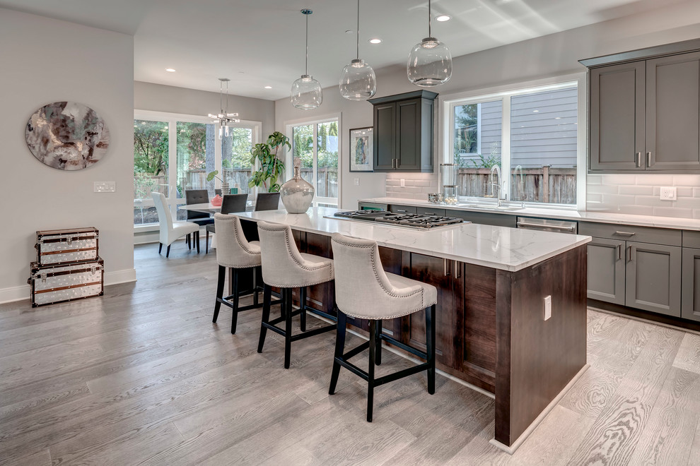 Large traditional l-shaped kitchen/diner in Seattle with recessed-panel cabinets, grey cabinets, engineered stone countertops, white splashback, ceramic splashback, stainless steel appliances, light hardwood flooring, an island, grey floors and white worktops.