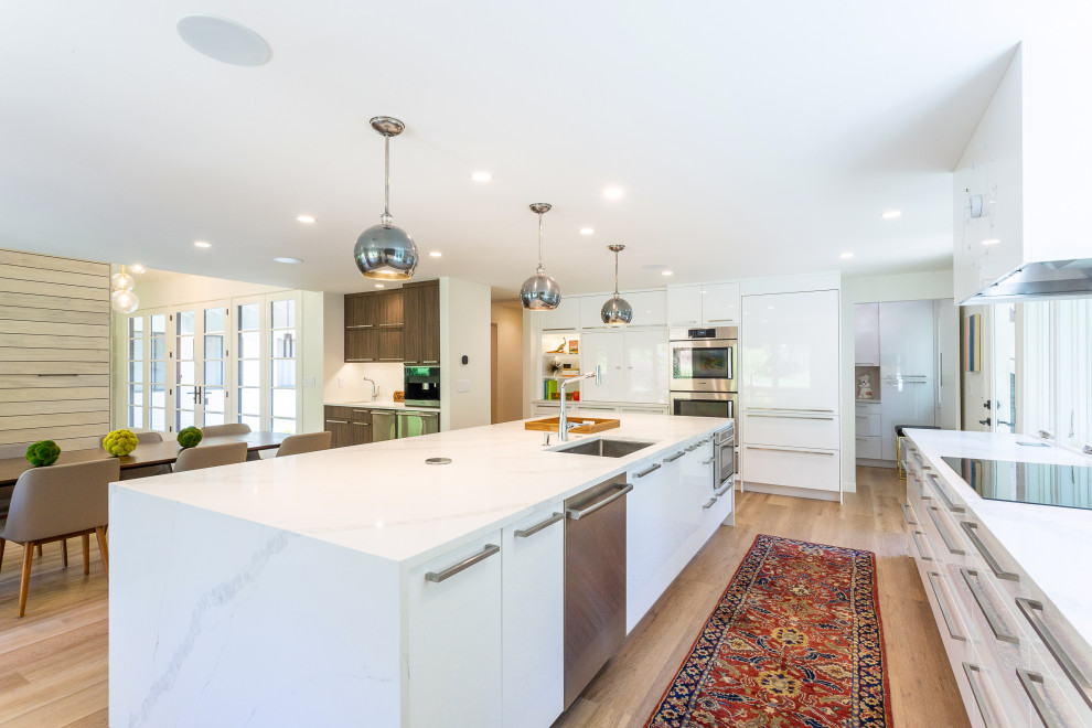 Modern inredning av ett stort vit linjärt vitt kök och matrum, med en undermonterad diskho, släta luckor, vita skåp, bänkskiva i kvartsit, vitt stänkskydd, stänkskydd i sten, integrerade vitvaror, ljust trägolv, en köksö och brunt golv