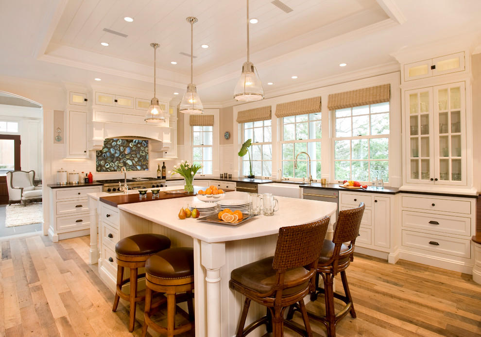 Inspiration for a coastal l-shaped medium tone wood floor kitchen remodel in Boston with a farmhouse sink, recessed-panel cabinets, white cabinets, white backsplash, stainless steel appliances and an island