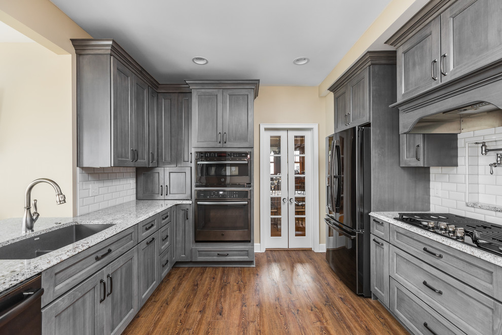 Ejemplo de cocina clásica renovada de tamaño medio con armarios con paneles empotrados, puertas de armario grises, salpicadero de azulejos tipo metro, electrodomésticos negros y una isla