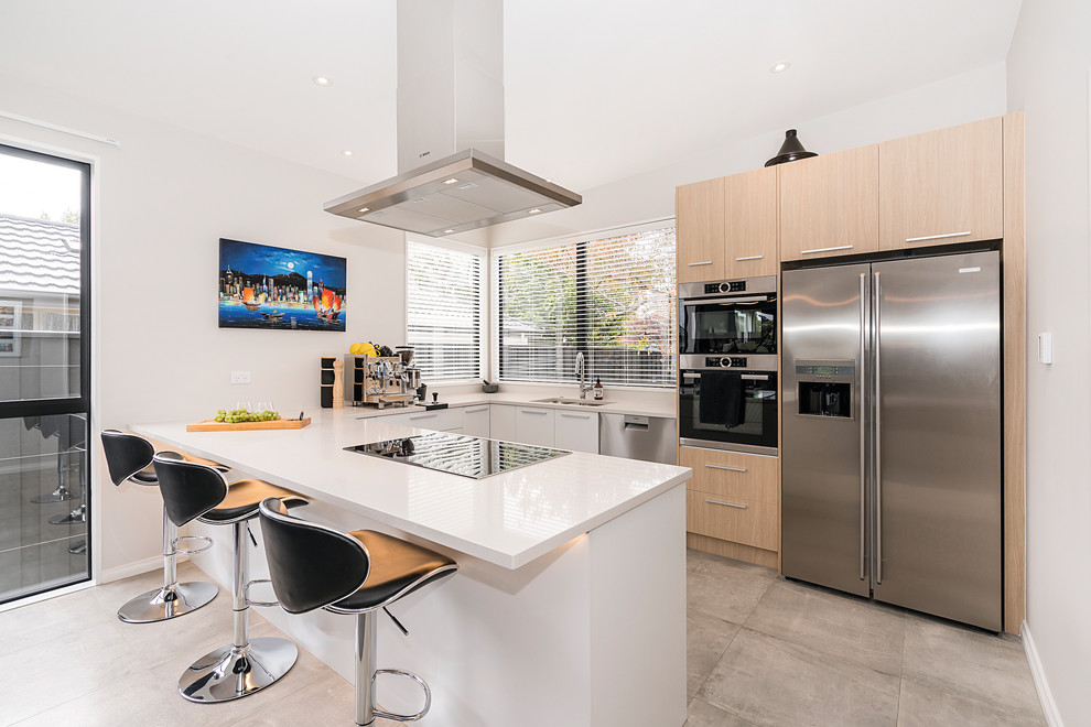 Example of a trendy u-shaped gray floor kitchen design in Christchurch with a double-bowl sink, flat-panel cabinets, light wood cabinets, stainless steel appliances, a peninsula and white countertops