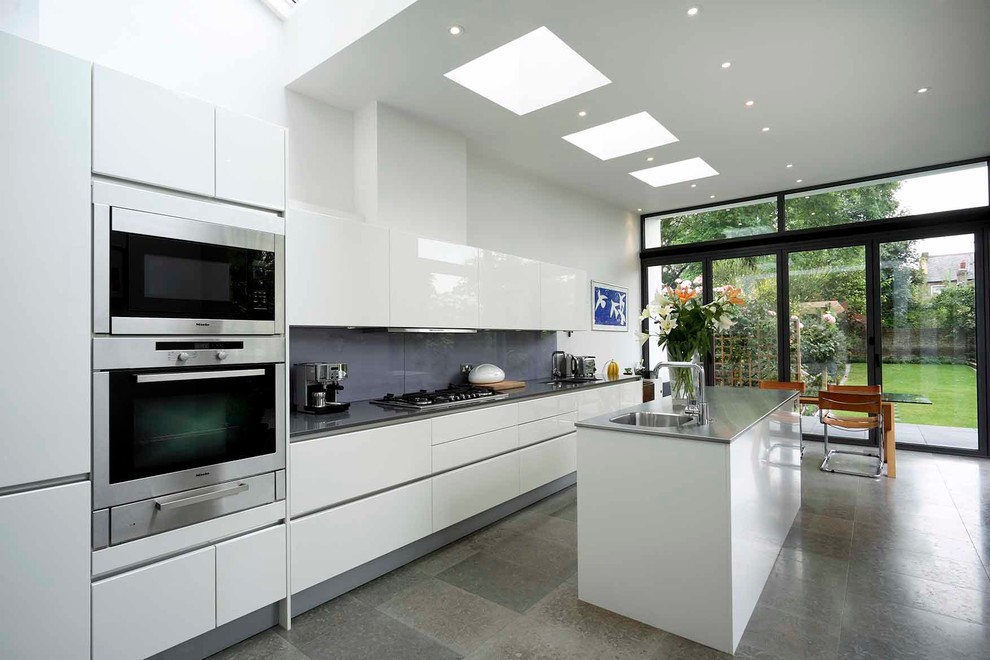 Contemporary kitchen/diner in London with a single-bowl sink, flat-panel cabinets, white cabinets, stainless steel worktops, grey splashback, glass sheet splashback, stainless steel appliances and an island.