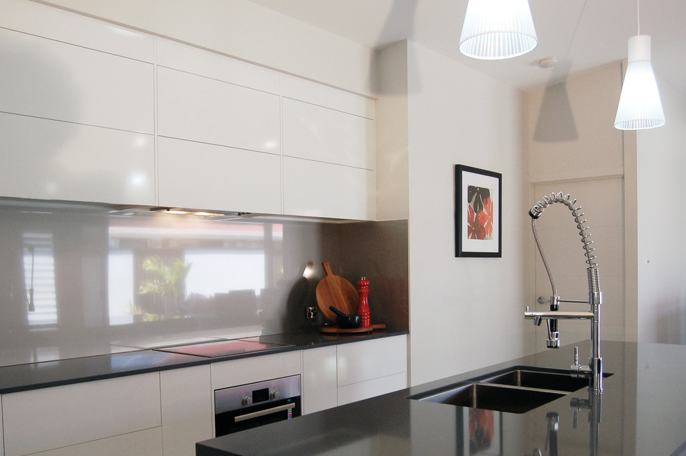 This is an example of a small contemporary galley open plan kitchen in Other with a submerged sink, flat-panel cabinets, white cabinets, composite countertops, glass sheet splashback, stainless steel appliances, ceramic flooring, an island, grey splashback, grey worktops and white floors.
