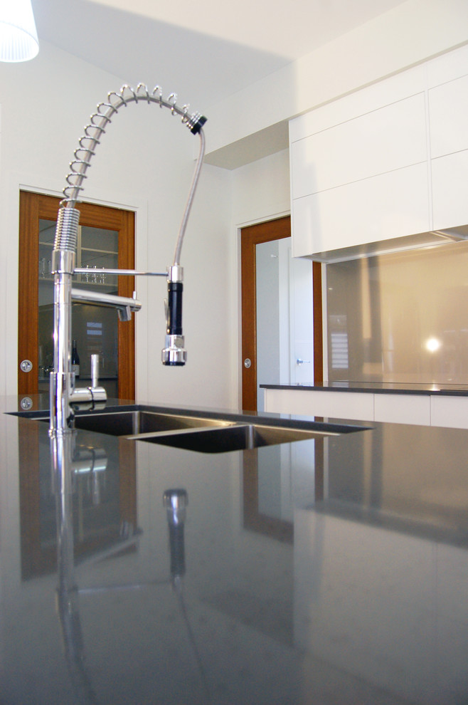 Photo of a small contemporary galley open plan kitchen in Other with a submerged sink, flat-panel cabinets, white cabinets, composite countertops, glass sheet splashback, stainless steel appliances, ceramic flooring, an island, grey splashback, white floors, grey worktops and a vaulted ceiling.